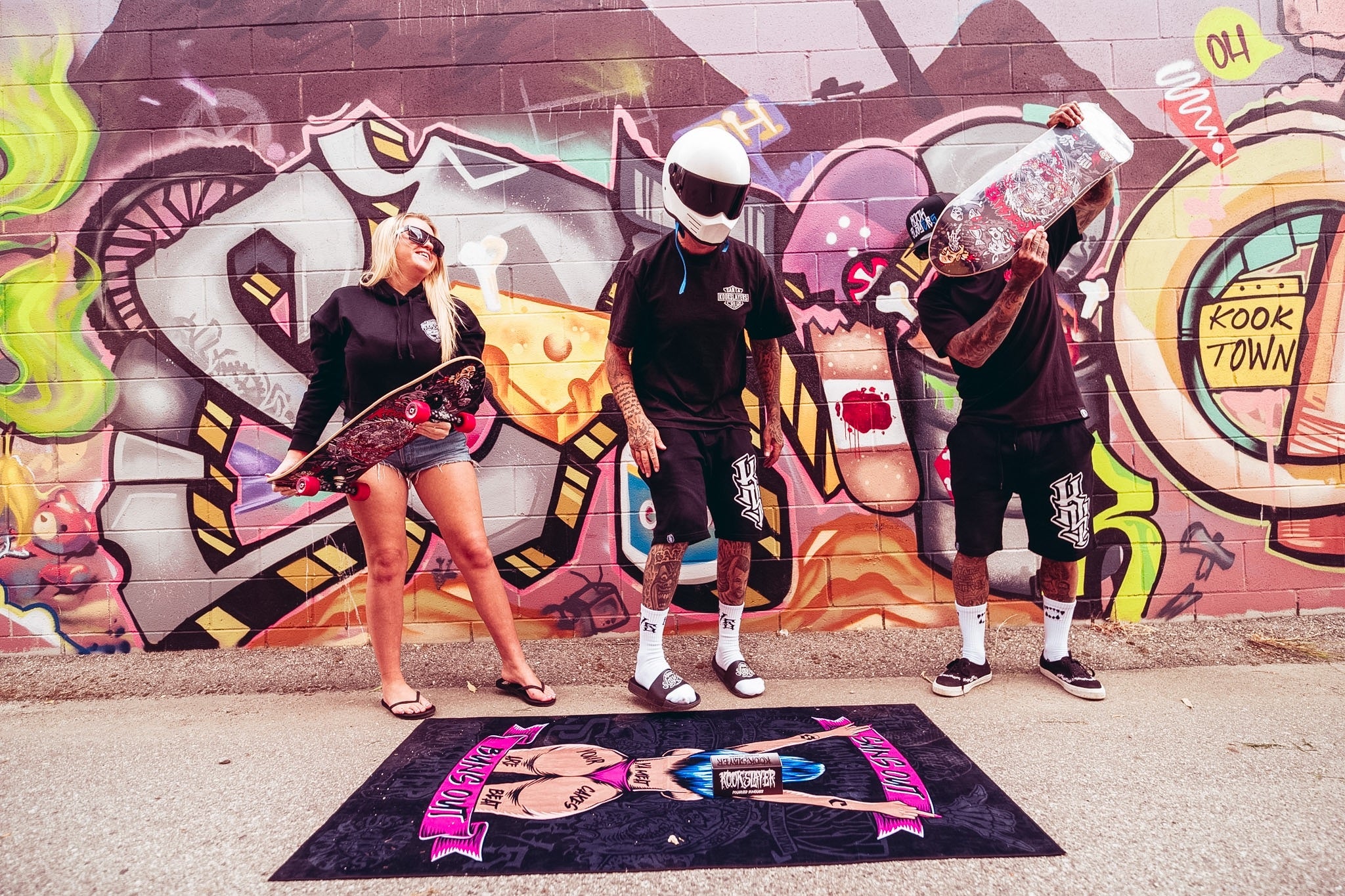 Three people in Kookslayer apparel with skateboards and an illustrated towel in front of a colorful mural