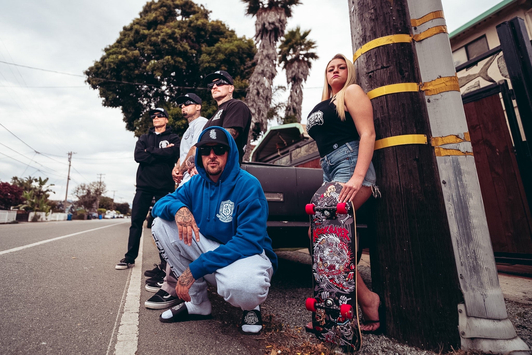 Kookslayer crew on a street beside a parked car, one crouched in a blue hoodie, one holding a skateboard