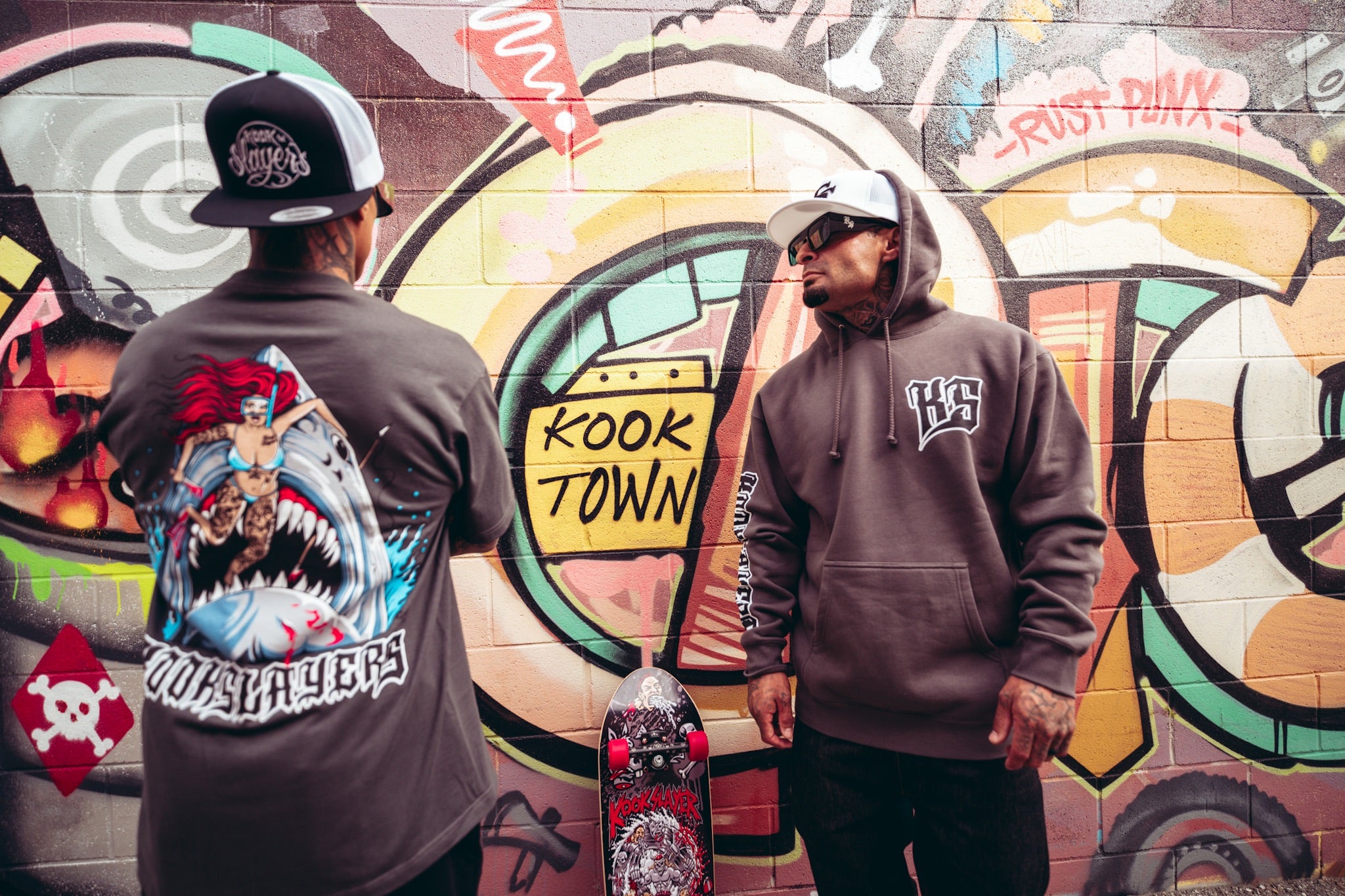 Back of the Colby Phillips Shark Tee next to a gray Kookslayer hoodie in front of the Kook Town mural